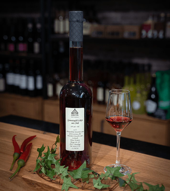 Eine Flasche mit rotem Likör und ein Weinglas mit rotem Wein stehen auf einem Holztheken. Dekoriert mit roten Chilischoten und grünen Efeublättern.